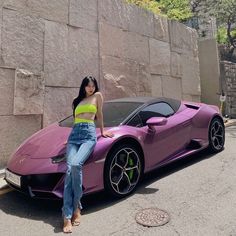 a woman sitting on the hood of a pink sports car in front of a stone wall