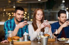 three people sitting at a table looking at their cell phones while eating food and drinking wine