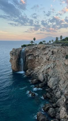 a waterfall on the side of a rocky cliff next to the ocean with palm trees in the background