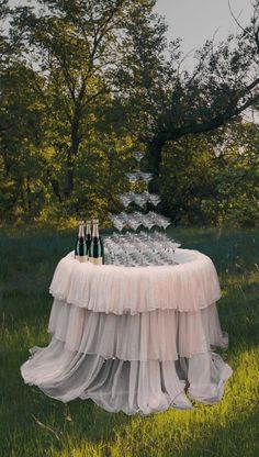 a table topped with bottles and glasses on top of a grass covered field next to trees