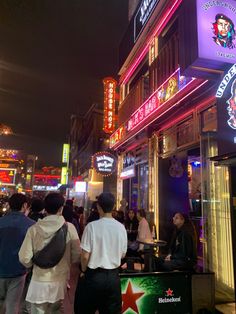 people are walking down the sidewalk in front of a building with neon signs on it