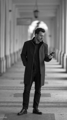 a man standing in an empty hallway looking at his cell phone while wearing a coat