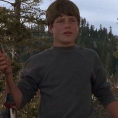 a young man holding a knife in his hand while standing next to a pine tree