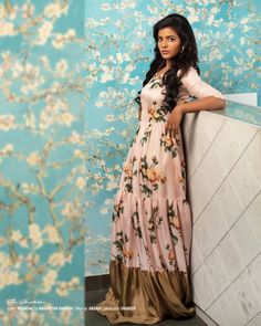 a woman in a long dress leaning against a wall with flowers on the wall behind her