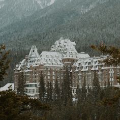 a large building surrounded by trees and snow covered mountains in the background with evergreens