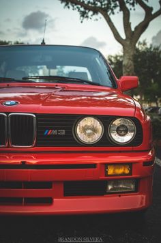 the front end of a red car parked in a parking lot next to a tree