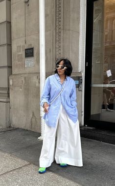 a woman standing on the sidewalk in front of a building wearing white pants and a blue shirt