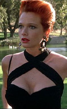 a woman with red hair wearing black dress and large earrings standing in front of water