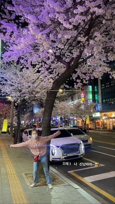 a woman standing next to a tree with purple flowers on it's branches in the city