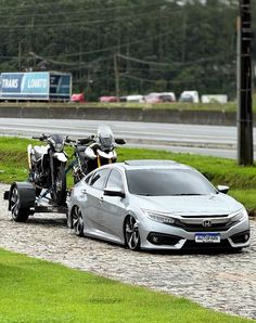 a motorcycle is attached to the back of a silver car on a road with grass and trees in the background