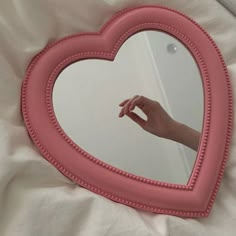 a pink heart shaped mirror on top of a white sheet with a hand reaching for it