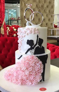 a wedding cake decorated with pink flowers and two rings on top, sitting on a round table