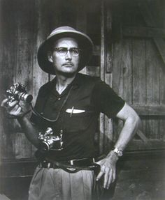 a black and white photo of a man wearing a hat holding a camera in his right hand