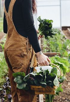 a woman in an apron holding a basket full of greens and carrots while standing next to a garden