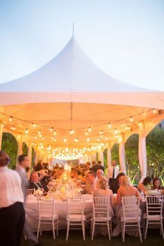 a group of people sitting at tables under a white tent with lights on the ceiling