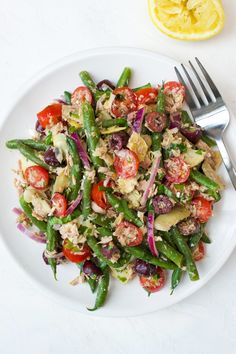 a white plate topped with green beans, tomatoes and other veggies next to a lemon wedge