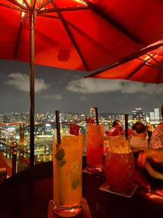 people are sitting under umbrellas at an outdoor bar with drinks on the table and city lights in the background