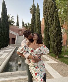 a woman in a floral dress kissing another woman's face near a fountain and trees