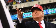 a man in a suit and red hat throwing a ball at a football game with an american flag on the sidelines