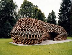 an unusual building made out of logs in the middle of a grassy area with trees behind it