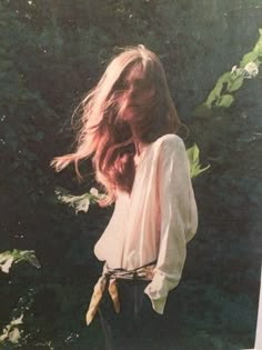 a woman standing in front of trees with her hair blowing in the wind, wearing a white blouse and black pants