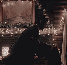 a man and woman sitting in front of a fireplace with christmas lights on the wall