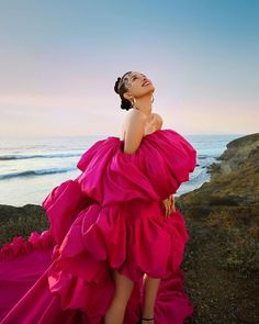 a woman in a pink dress is standing on the rocks near the ocean and looking up into the sky