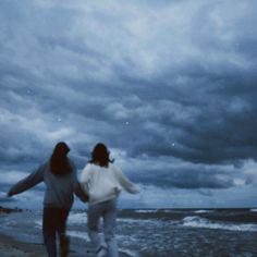 two women are walking along the beach holding hands and looking at the ocean under a cloudy sky