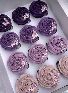 twelve cupcakes with purple frosting and pearls on them in a white box