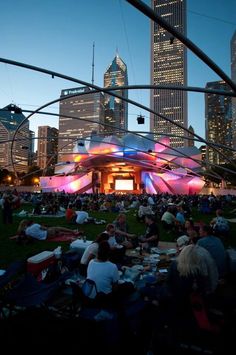 people are sitting on the grass in front of an outdoor movie screen at dusk with city buildings in the background