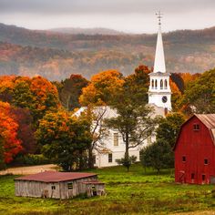 an old barn sits in the foreground with a church steeple and autumn foliage behind it