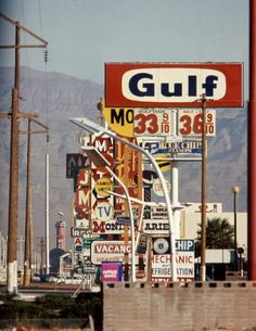 a gas station sign with many signs on the side of it and mountains in the background