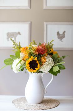 a white vase filled with lots of flowers on top of a table next to framed pictures