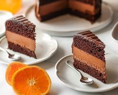 three slices of chocolate cake on white plates with oranges and spoons next to them