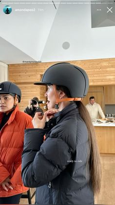 two women wearing helmets are standing in a kitchen and one is looking at her phone