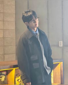 a young man standing in front of a book shelf wearing glasses and a black coat