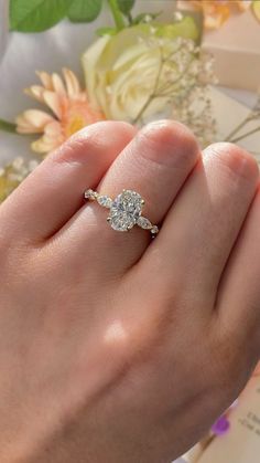 a woman's hand with a diamond ring on top of her finger and flowers in the background