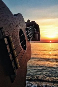 an acoustic guitar sitting on top of a wooden table next to the ocean at sunset