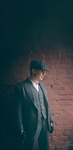 a man standing in front of a brick wall wearing a suit and hat with one hand on his hip