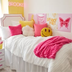 a white bed topped with lots of pink and yellow pillows next to a wall covered in pictures