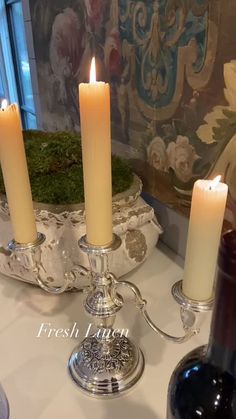 three candles are lit on a table next to some wine bottles and a vase with moss in it