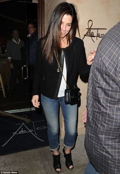 a woman in jeans and a black jacket is walking out of a building with her hand on her hip