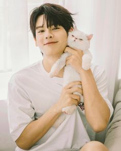 a young man holding a white cat in his hands and smiling at the camera while sitting on a couch