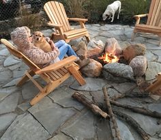 an elderly woman sitting in a chair next to a fire pit with two dogs on the other side