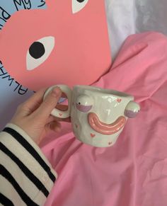 a hand holding a coffee mug with a face on it next to a pink teddy bear