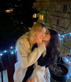 two women standing next to each other on a balcony at night with lights in the background