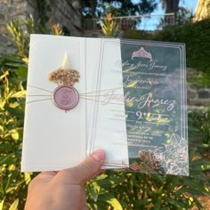 a person holding up two wedding cards in front of some plants and bushes, one with a wax stamp on it