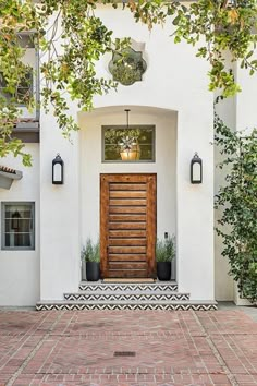 a white house with a wooden door surrounded by greenery and potted plants on the front porch