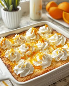 an orange dessert with whipped cream on top in a white casserole dish next to fresh oranges