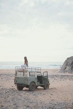 a woman standing on top of a green jeep in the sand next to water and rocks